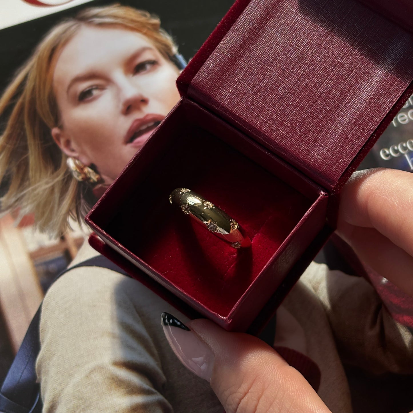 Gold ring in a red box held by a hand, with a magazine featuring a woman's face in the background.
