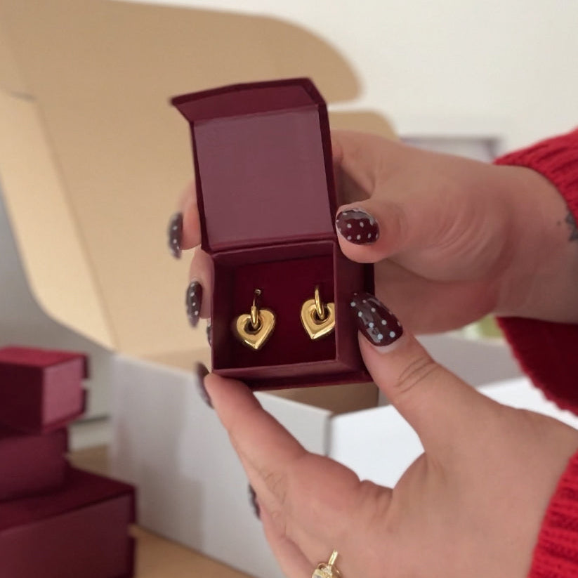 Gold heart-shaped earrings in a red jewellry box held by a person with red nail polish.