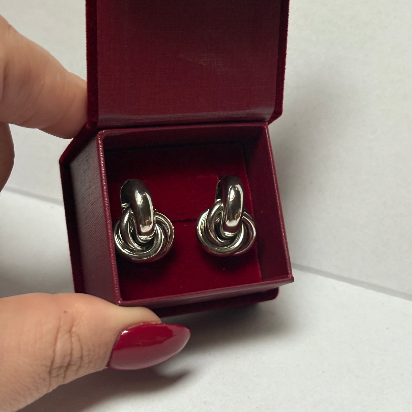 Silver knot-shaped cufflinks in a red box held by a hand with red nail polish.