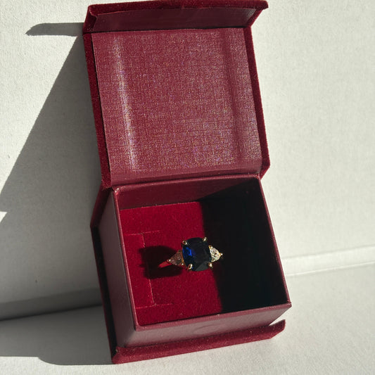 Open red jewelry box with a ring featuring a blue gemstone on a light gray background
