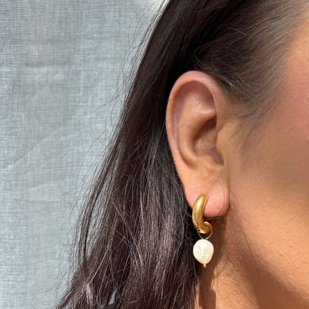 Close-up of an ear wearing a gold hoop earring with a pearl charm against a light gray background.

