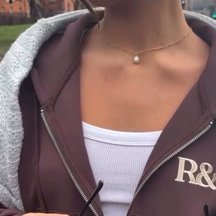 Person wearing a brown tracksuit with 'R&' logo, holding sunglasses outdoors.

