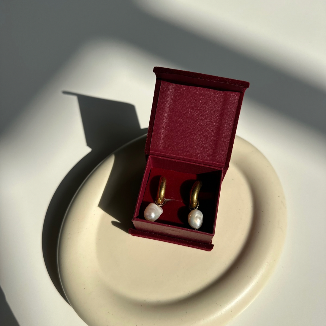 Pair of pearl earrings in a red jewelry box on a reflective surface with soft shadows.